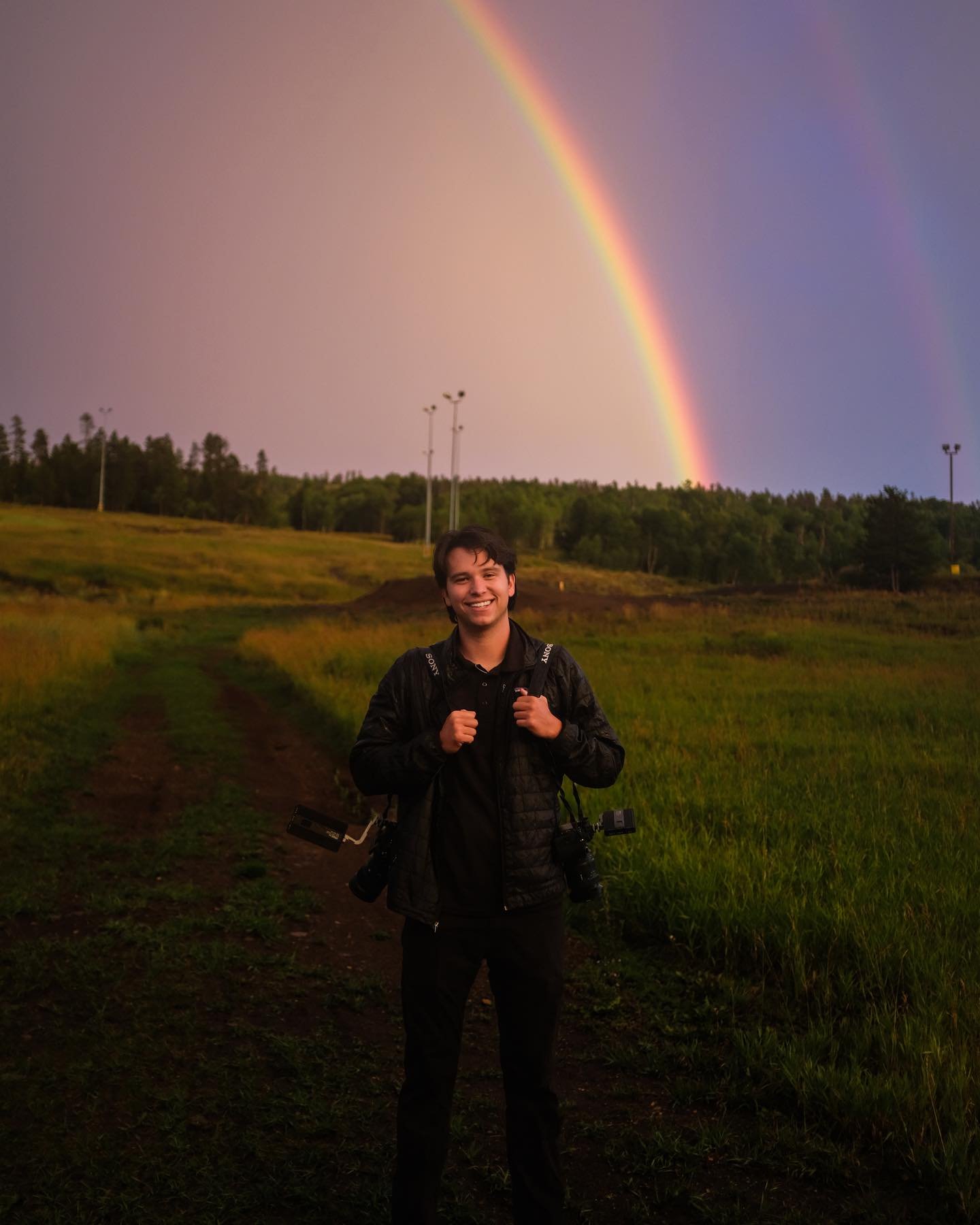 Matthew Weathersby, wedding filmmaker, standing in a field under a rainbow with his cameras
